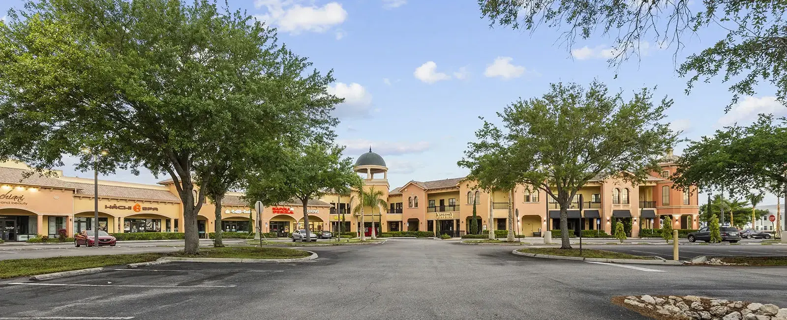 Front view of Sugden Park Plaza in Naples, Florida, featuring the commercial center's storefronts, customer entrances, covered walkways, landscaping, and parking area.