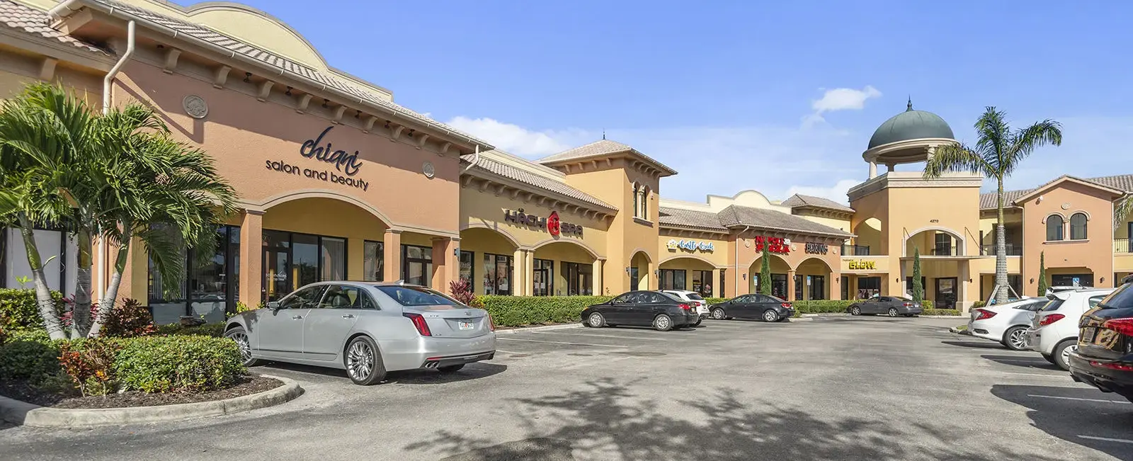 Left-side exterior view of Sugden Park Plaza in Naples, Florida, showing a portion of the commercial building with storefronts, covered walkways, architectural details, and landscaped areas.