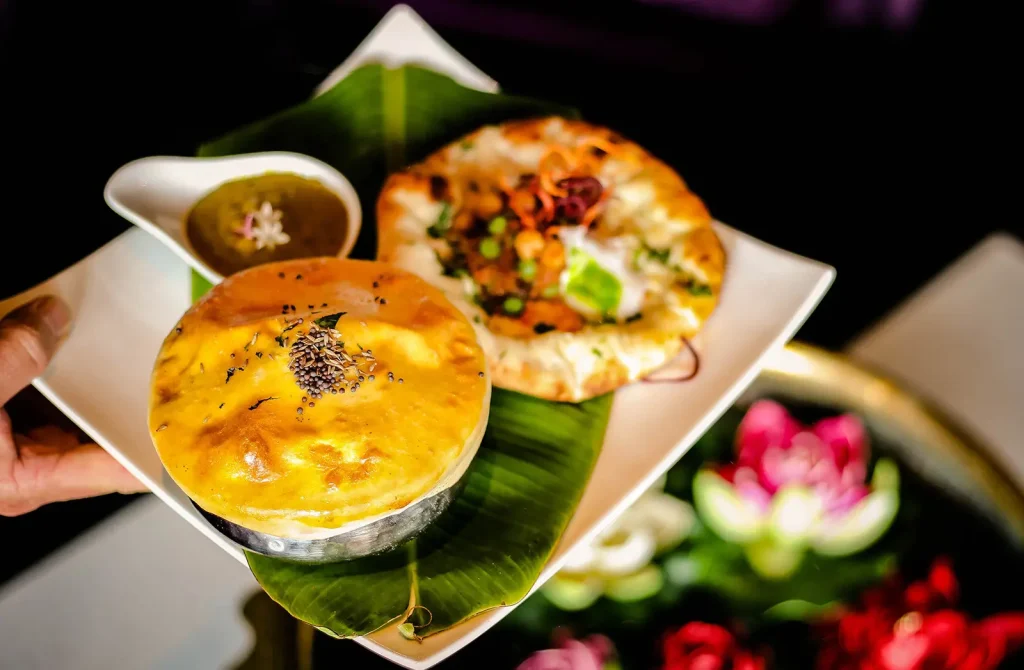 Golden-brown baked Indian dish served in a small metal bowl alongside a colorful naan topped with vegetables and sauce, presented on a white plate lined with banana leaves.